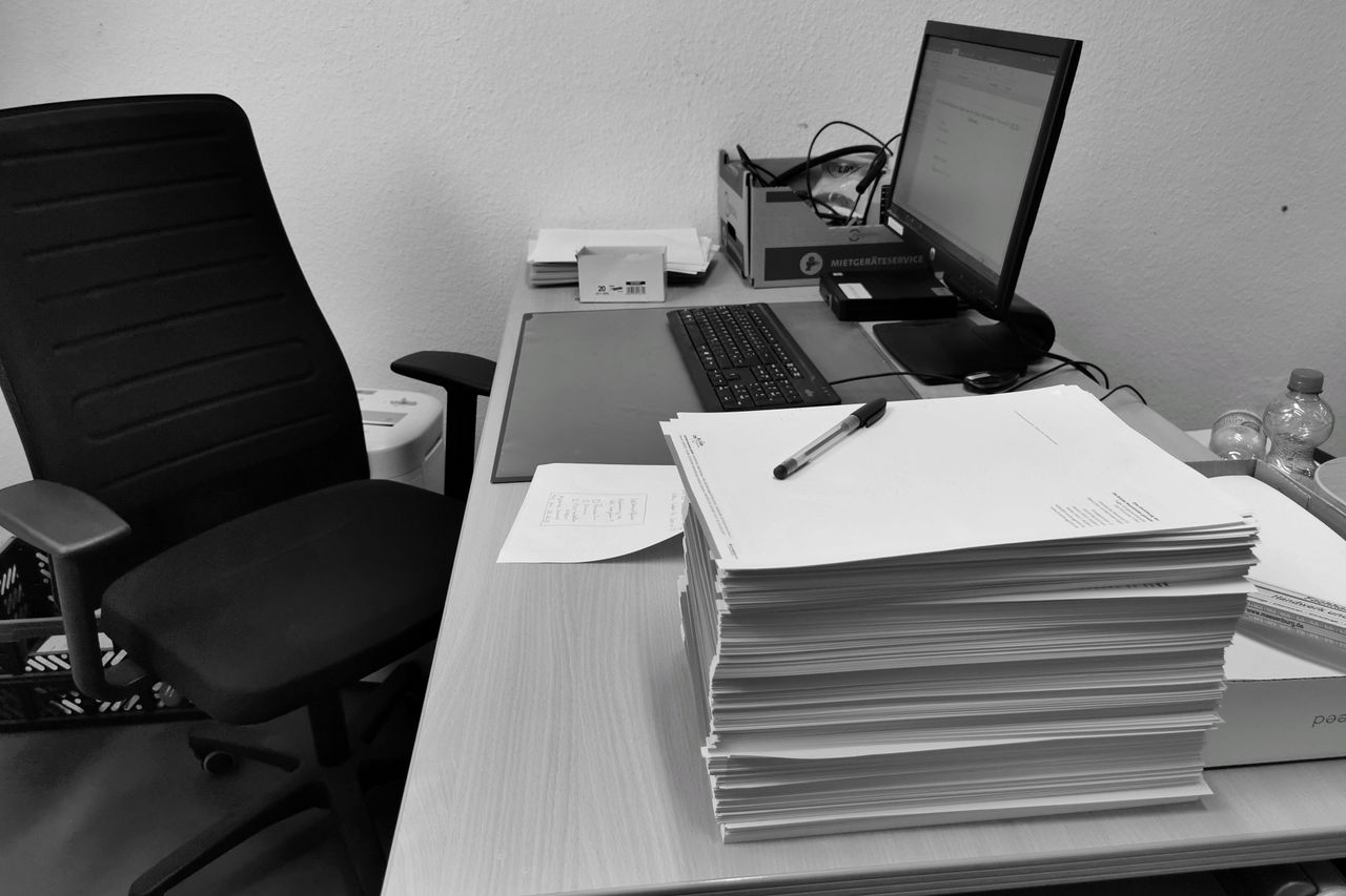 Accueil Black and white image of a cluttered office desk with a computer, chair, and paperwork.