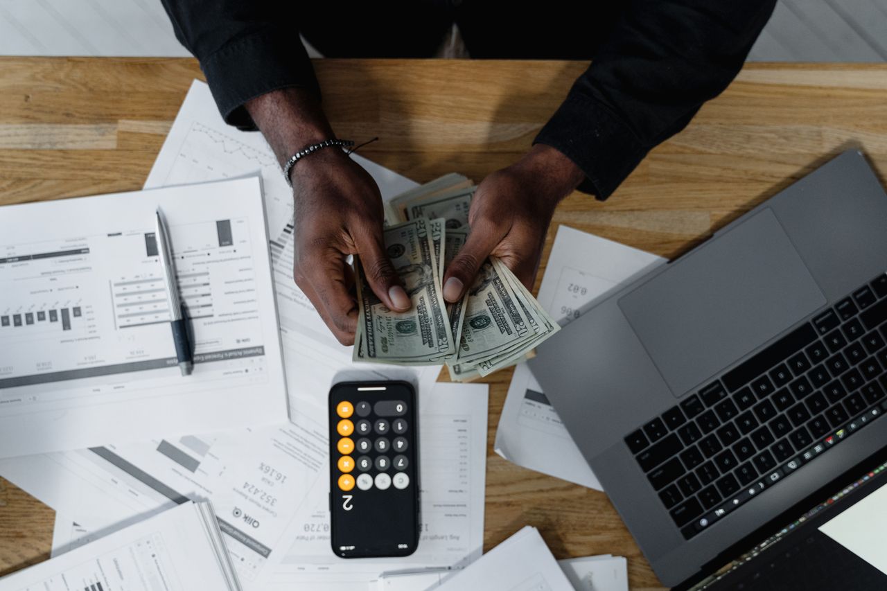 Accueil Person counting US dollars, using a calculator and laptop, with financial documents on a wooden desk.