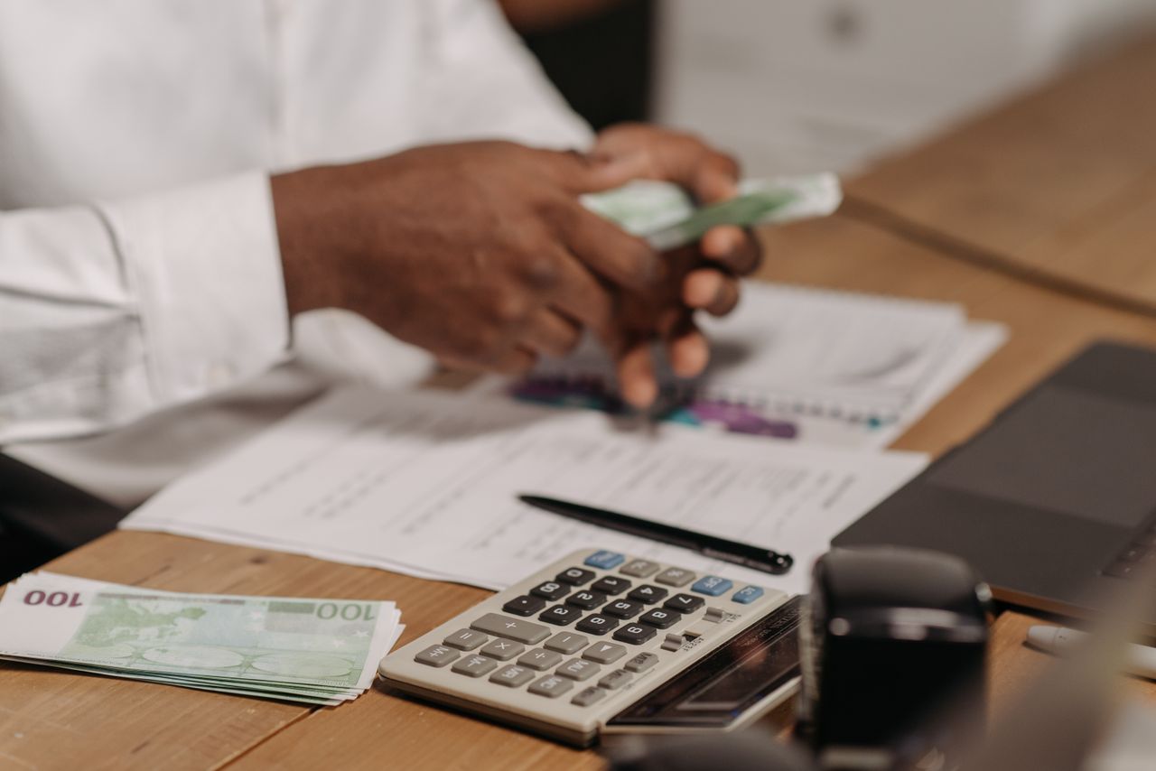 Accueil Hands counting euro bills on a wooden desk with calculator, financial documents, and laptop nearby.